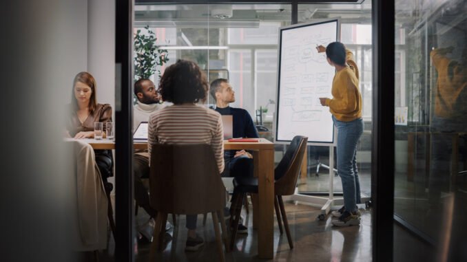 Project Manager Makes a Presentation for a Young Diverse Creative Team in Meeting Room in an Agency. Colleagues Sit Behind Conference Table and Discuss Business Development, User Interface and Design