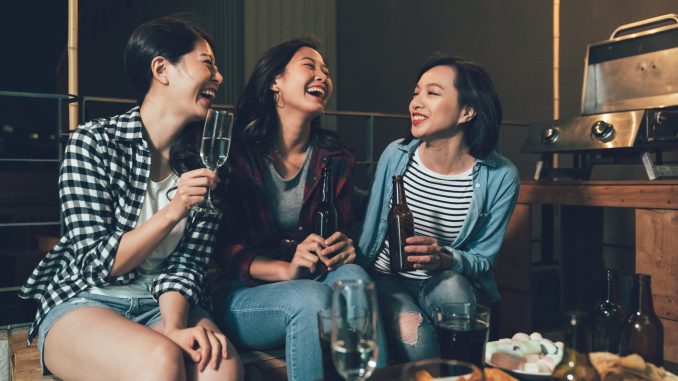 group of young people in rooftop party talking sitting on wooden chairs. happy girls enjoy barbecue at night on the roof. full of alcohol drinks and food on the table outdoors.