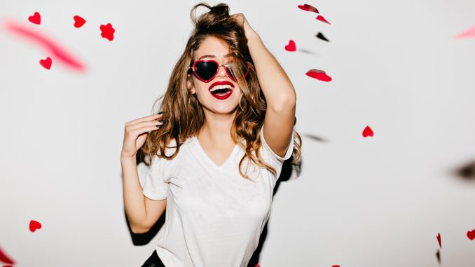 Indoor portrait of amazing caucasian female model in trendy t-shirt touching her long shiny hair. Laughing refined woman in sunglasses having fun with red confetti.