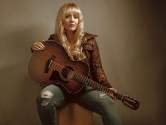 young blond woman sitting on a chair wearing blue jeans and brown jacket playing the guitar
