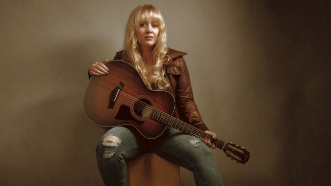 young blond woman sitting on a chair wearing blue jeans and brown jacket playing the guitar