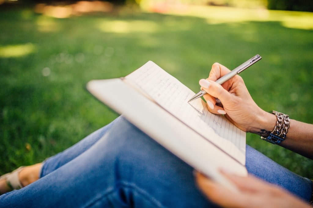women journaling sitting on green grass holding a pen