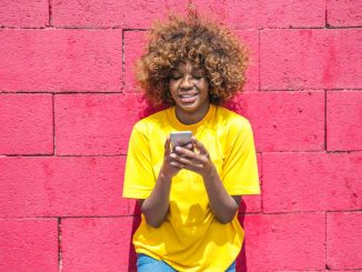 woman wearing a yellow t shirt, against pink wall, looking at the phone