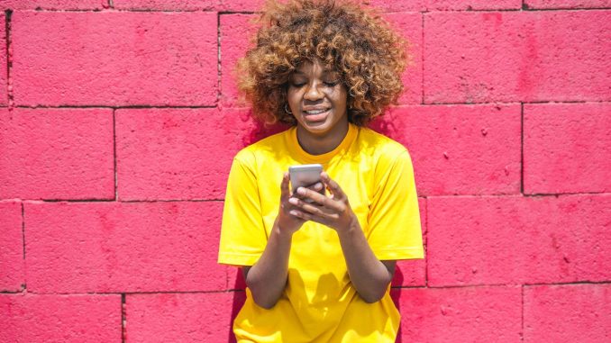 woman wearing a yellow t shirt, against pink wall, looking at the phone