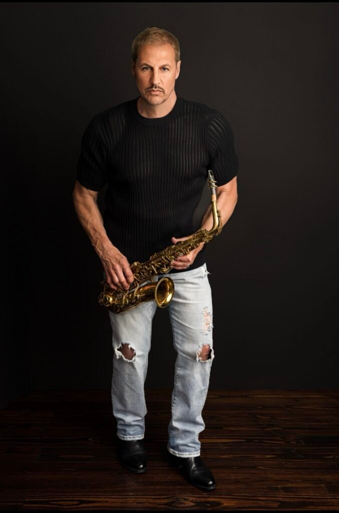 Portrait of saxophonist Michael Cates holding a saxophone in a black shirt and ripped jeans