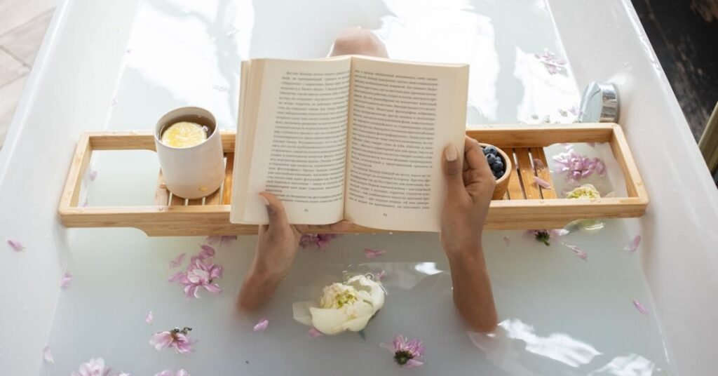 Person reading a psychology book in a relaxing flower bath with tea and fruit on a wooden tray.