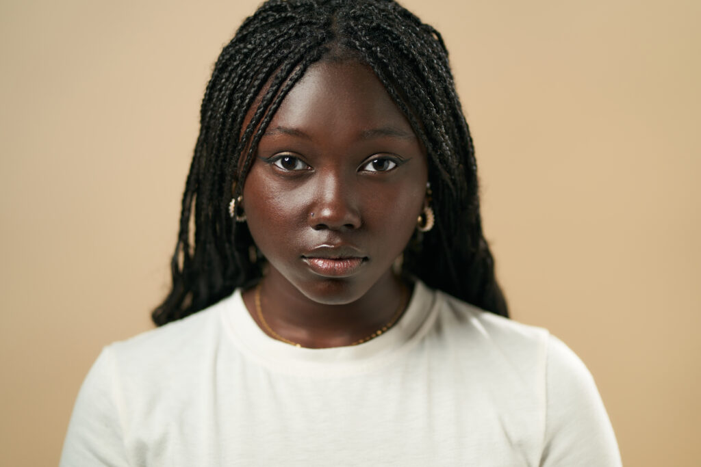 Studio portrait of a young black woman with long braids wearing a white t-shirt, looking directly at the camera with a serious expression