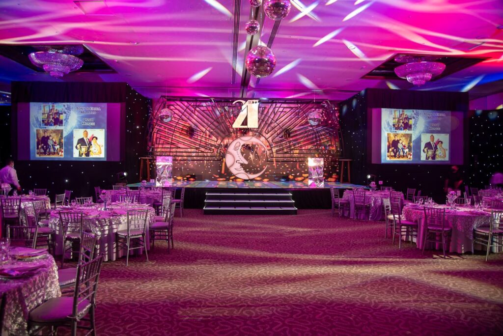 Corporate event venue with stage, banquet tables, colorful lighting, and presentation screens for a company gala or awards ceremony.